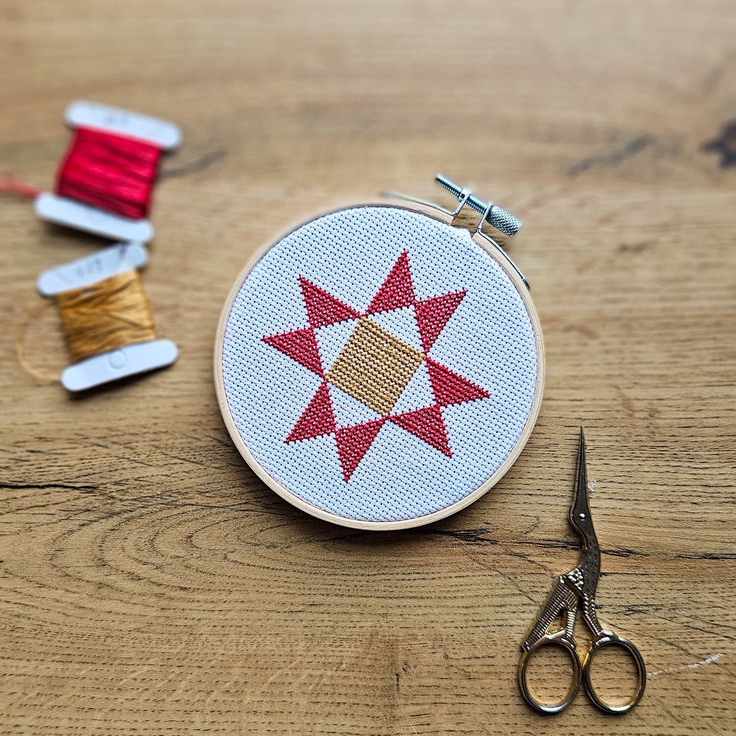 A round embroidery hoop holding white fabric with a red geometric Ohio star design and a tan center, styled on a wooden surface with spools of thread and scissors nearby.