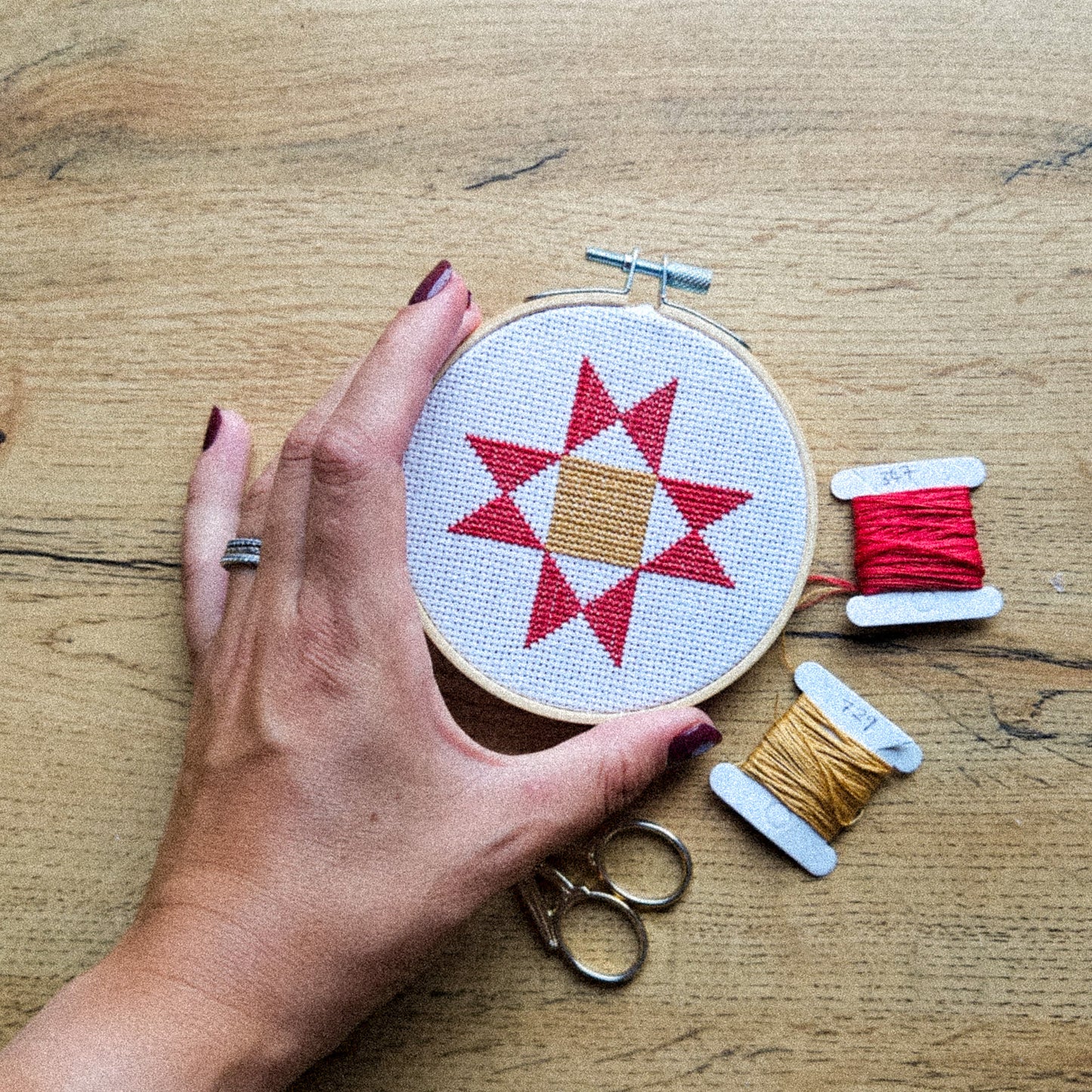 A hand holds a wooden embroidery hoop with a cross-stitch pattern of a red star and tan center on white fabric, set on a wooden table with spools of red and tan thread and metal scissors.