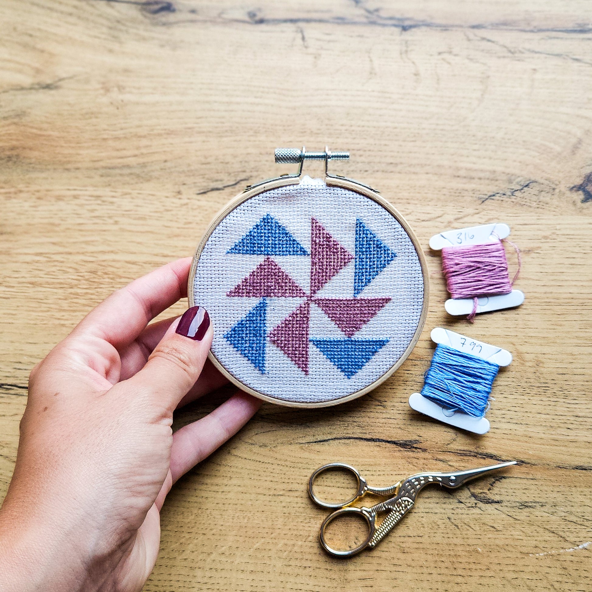 A hand with dark red nail polish holds a round embroidery hoop containing a geometric pinwheel flying geese pattern in blue and dusty pink thread on white fabric. The scene is set on a light wood table alongside two spools of thread in pink and blue, and a pair of silver embroidery scissors.