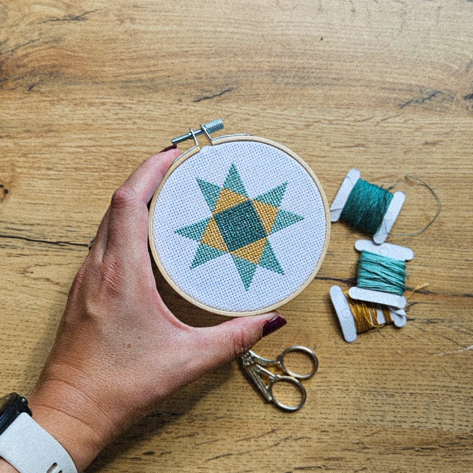 A hand holds a wooden embroidery hoop with a completed cross-stitch pattern of a geometric star in teal and mustard yellow on white fabric, set on a wooden tabletop with spools of thread and scissors nearby.