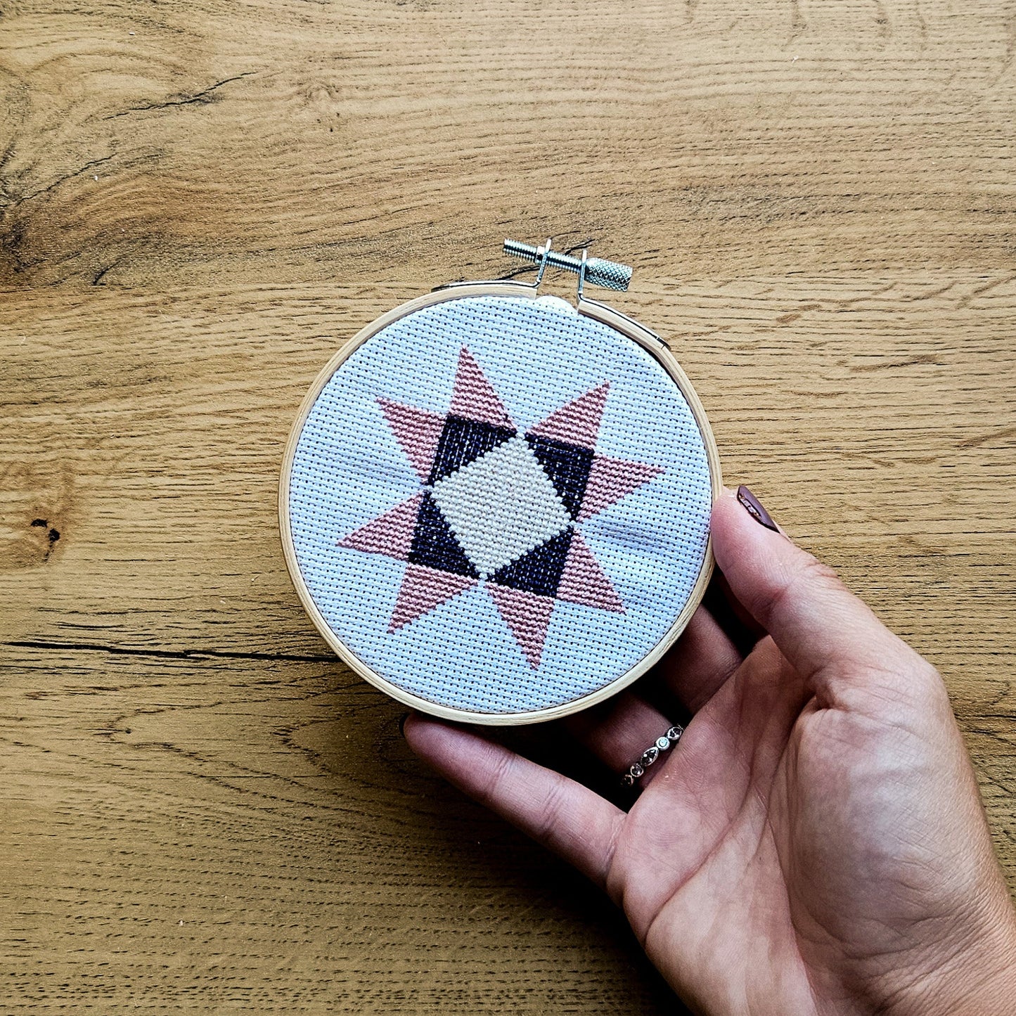 A hand holding a small wooden embroidery hoop with a finished geometric star cross-stitch design in pink, black, and cream on white fabric, set against a rustic wooden tabletop.