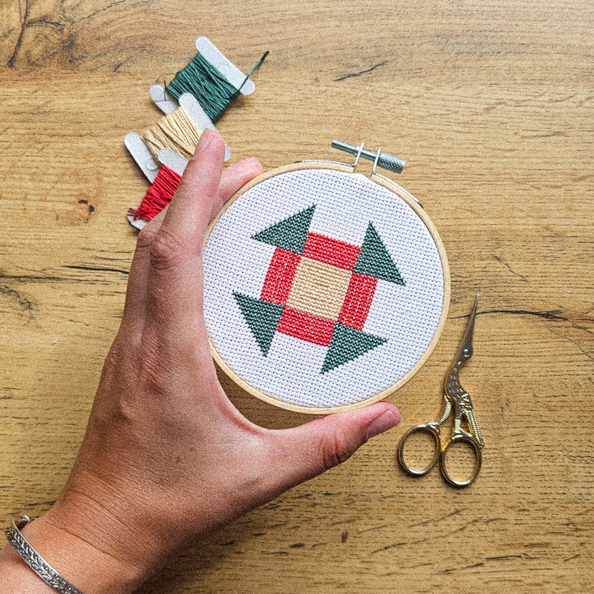 A hand holds a wooden embroidery hoop containing a cross-stitch pattern of green and red geometric shapes on white thread, set on a wooden table with spools of thread and scissors nearby.