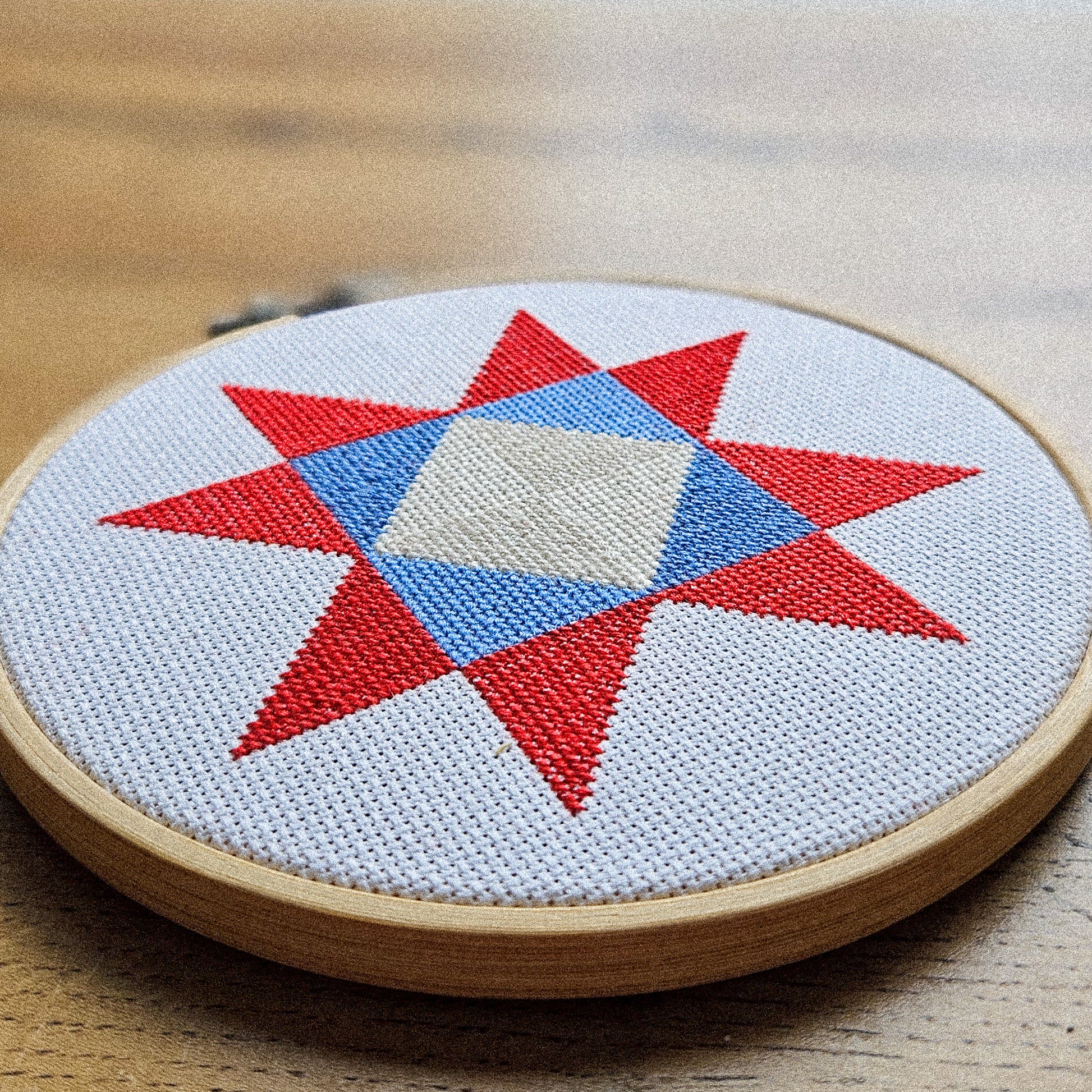 A circular embroidery hoop on a wooden table, featuring a completed cross-stitch design of a red and blue star with a white center on white fabric.