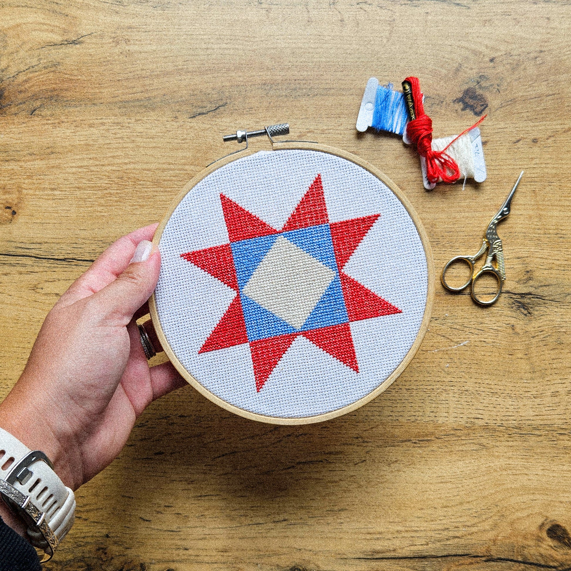 A hand holds a wooden embroidery hoop containing a completed cross-stitch pattern of a red and blue star on white fabric, set on a wooden tabletop with embroidery floss and scissors nearby.