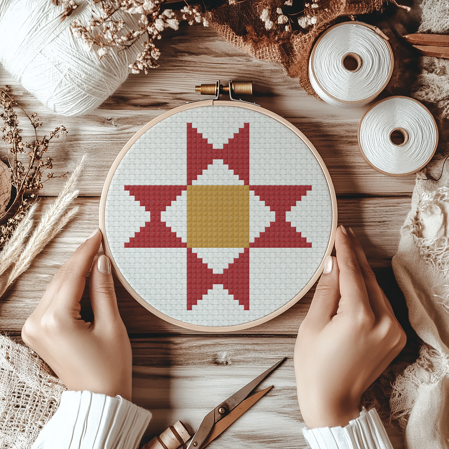 A top-down view of a person's hands holding a circular embroidery hoop with a cross-stitch pattern of a star and diamond. The scene is styled with spools of white thread, yarn, dried flowers, and a wooden surface.