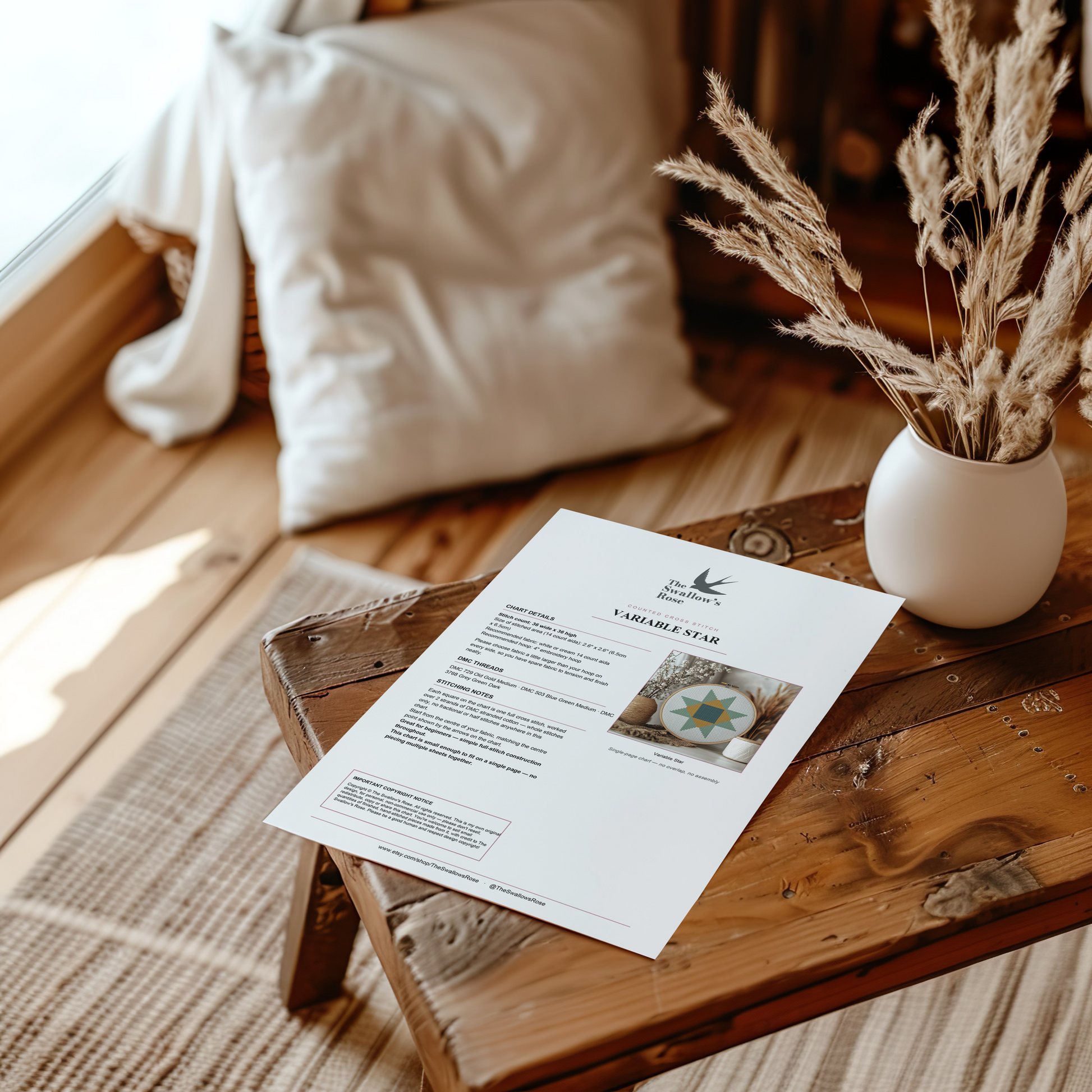 A printed sewing pattern sheet for a 'VARIABLE STAR' design lies on a rustic wooden table in a cozy, sunlit room. The scene is styled with a white pillow, a small white vase containing dried grasses, and a woven rug on a wooden floor.