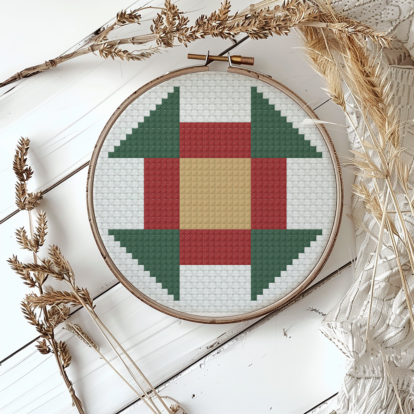 A circular embroidery hoop displaying a geometric cross-stitch pattern with green, red, beige, and white blocks, styled on a white wooden surface with dried wheat and lace.