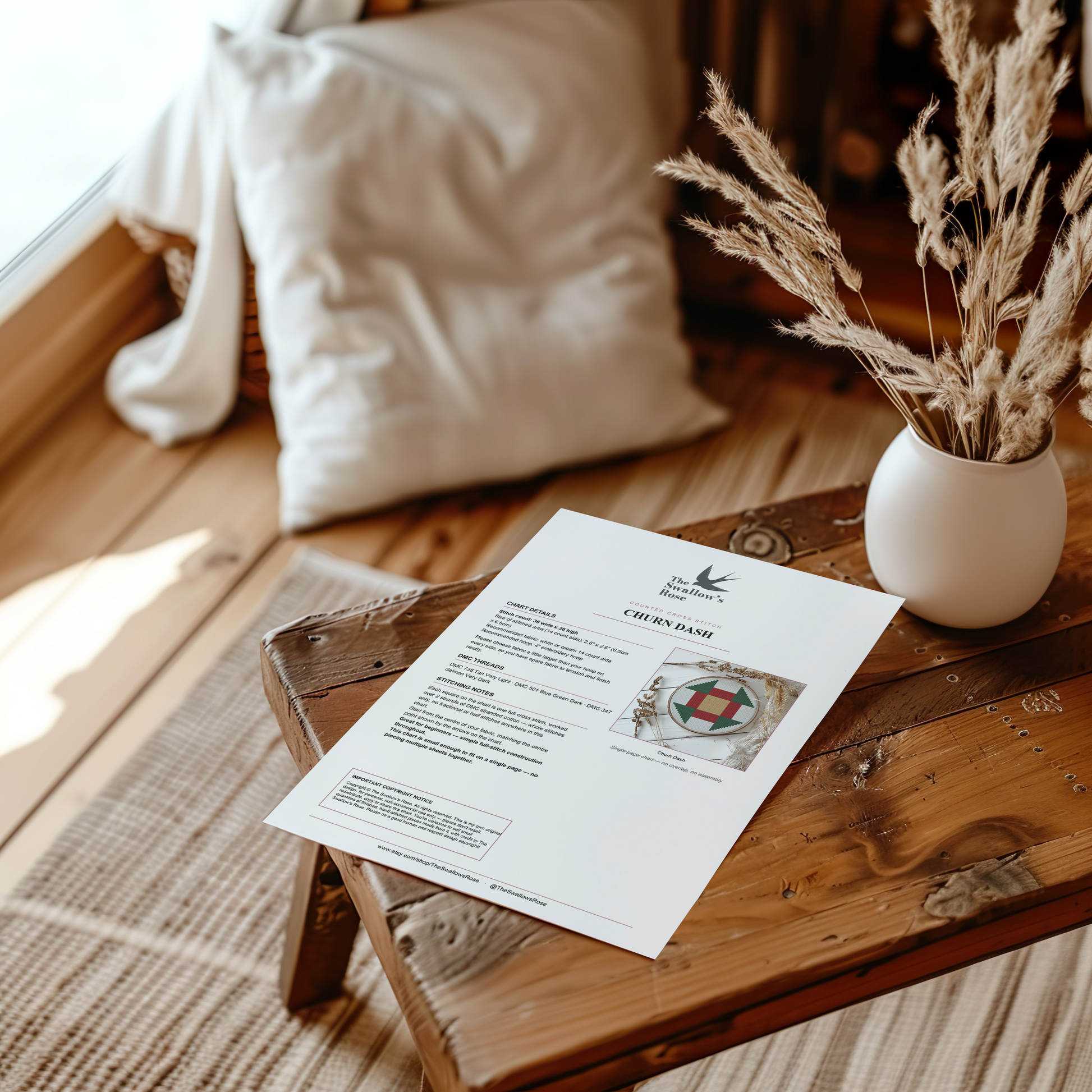 A printed white card with the title 'CHURN DASH' and a decorative emblem lies on a rustic wooden table in a warm, softly lit room. A white vase with dried grasses and a cream pillow are blurred in the background.