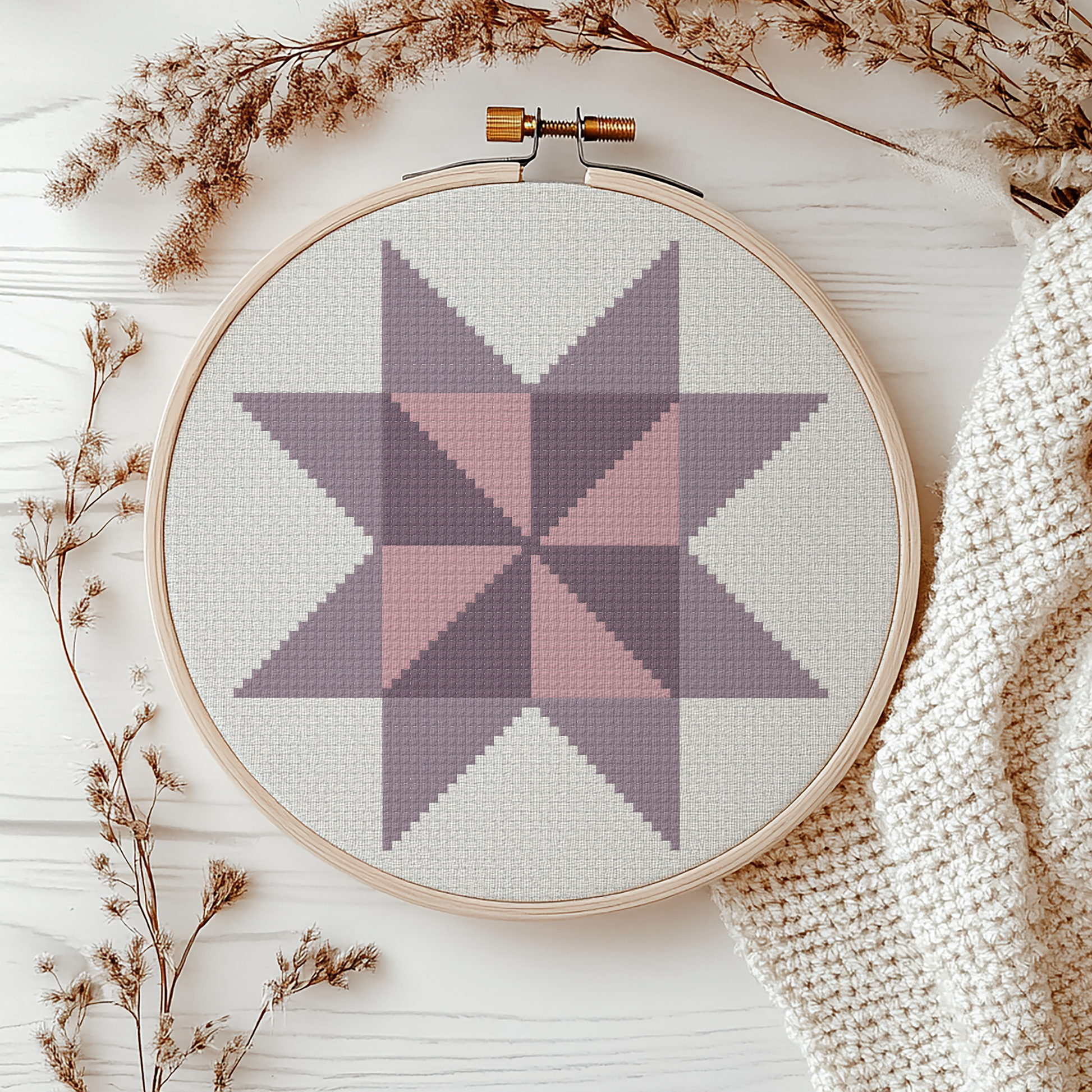A circular embroidery hoop holding light gray fabric with a geometric star pattern in muted pink and lavender-gray tones, styled on a white wood surface with dried flowers and a cream knit blanket.