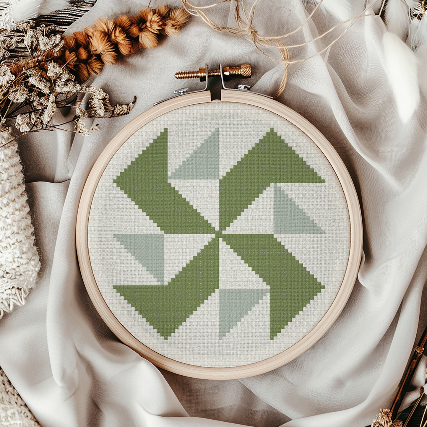 A circular embroidery hoop holding a geometric cross-stitch design in shades of sage green and light gray on white thread, styled with draped cream fabric, dried flowers, and a knit textile.