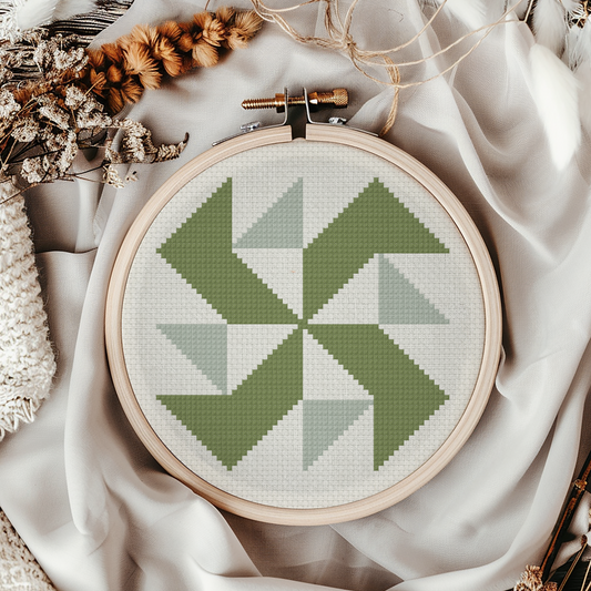 A circular embroidery hoop holding a geometric cross-stitch design in shades of sage green and light gray on white thread, styled with draped cream fabric, dried flowers, and a knit textile.