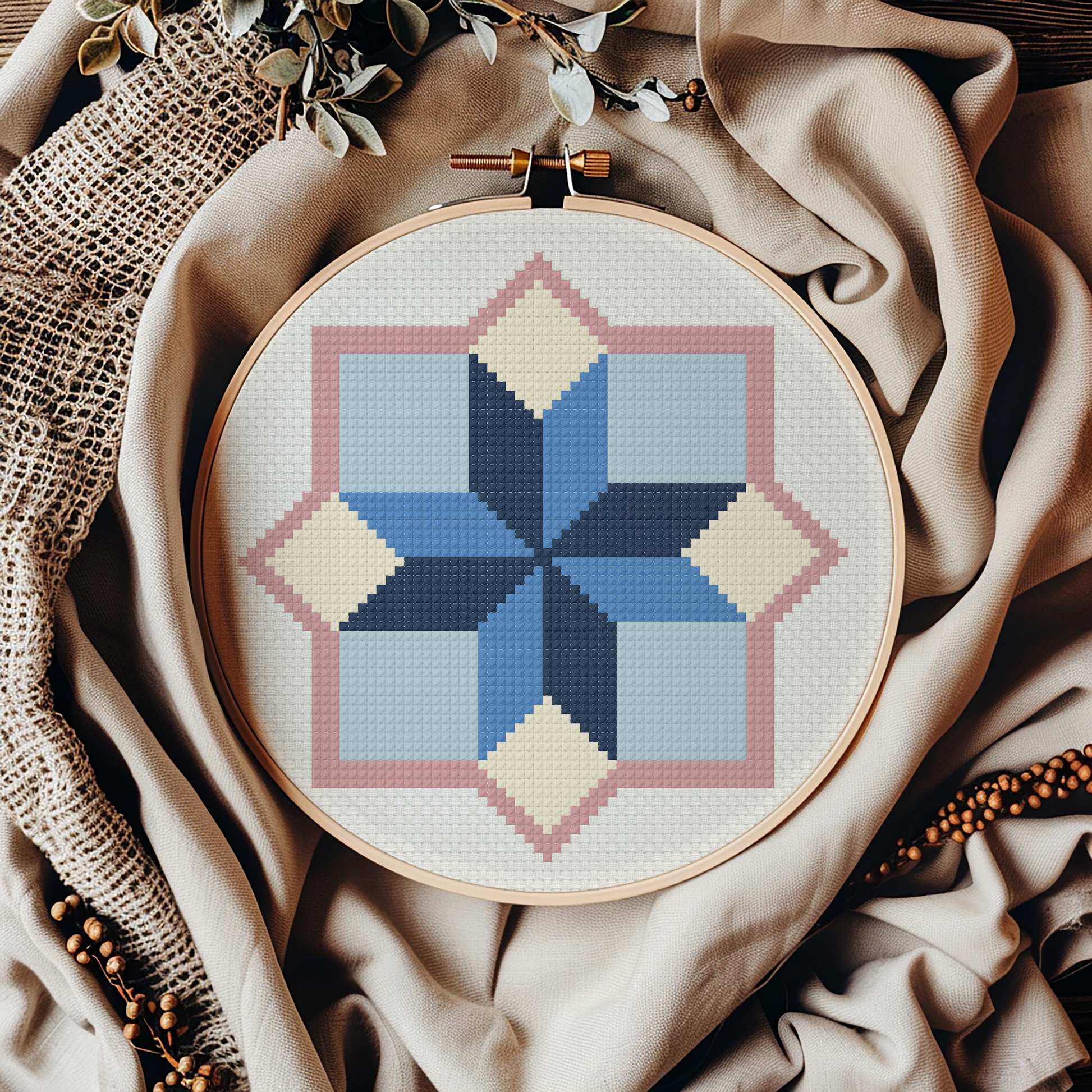 A circular embroidery hoop holding a geometric cross-stitch pattern in shades of blue, navy, pink, and cream, styled on draped beige fabric with burlap, beads, and dried leaves.