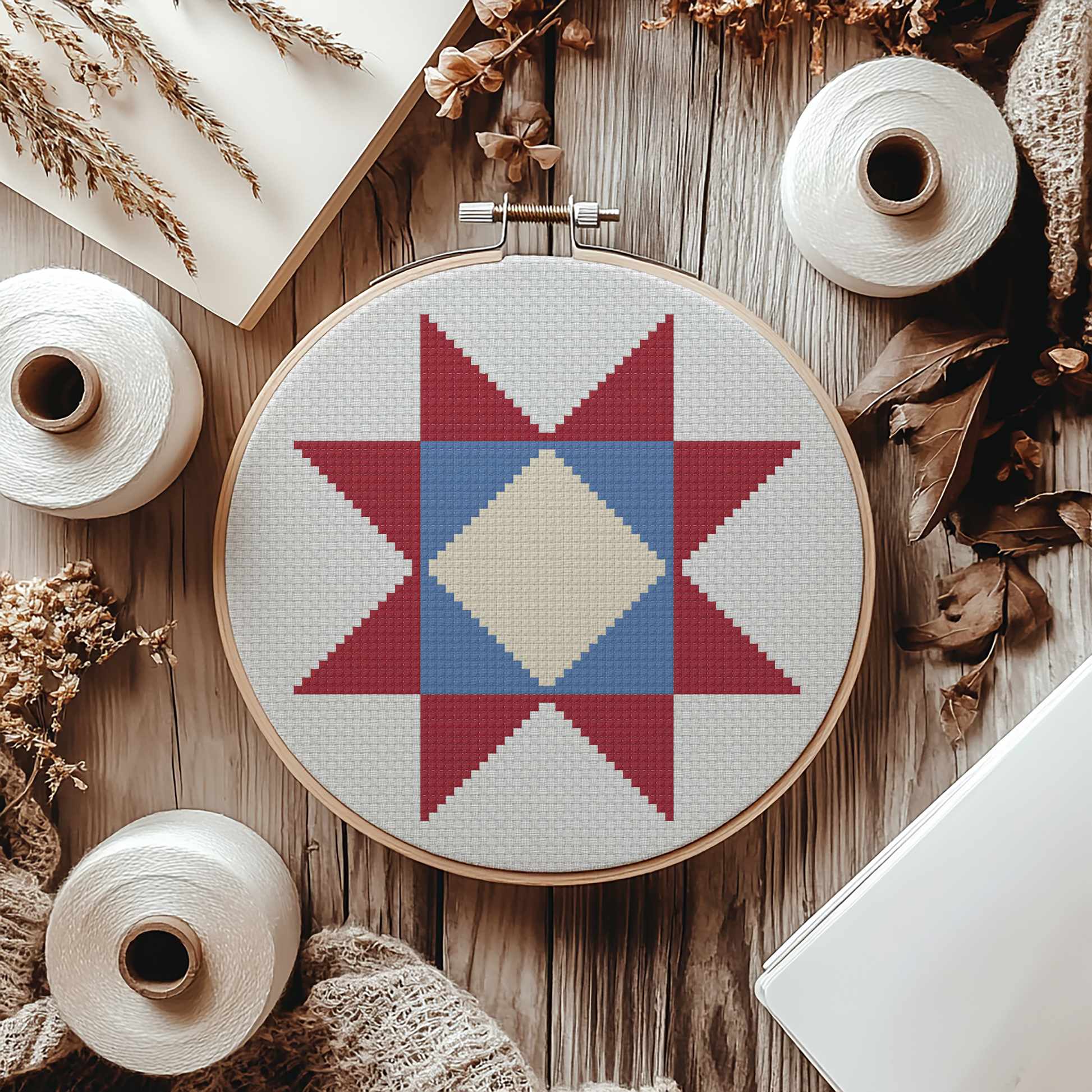 A cross-stitch pattern displayed in a wooden embroidery hoop, featuring a geometric star design in red, blue, and cream on a white fabric. The hoop is styled in a flat-lay arrangement on a rustic wooden table, surrounded by spools of white thread, dried flowers, and a notebook.